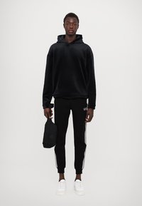 Felpa nera in pile, pantaloni sportivi neri a gamba stretta con strisce laterali, sneakers bianche, che tiene in mano una piccola borsa nera, su uno sfondo neutro.