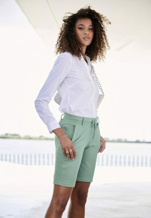 Shorts vert clair avec taille élastique et cordon de serrage, associés à une chemise blanche ajustée à boutons. Tissu lisse et design cintré.