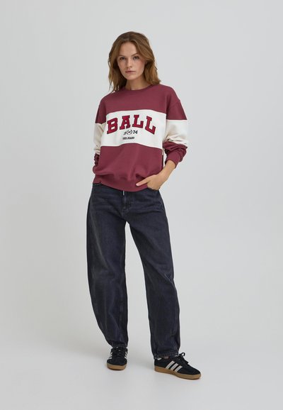 Felpa maroon e bianca con la scritta "BALL 74", abbinata a jeans loose-fit blu scuro e scarpe nere con strisce bianche. Tessuto dalla texture morbida.