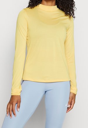 Kvinde iført en gul langærmet tætsiddende top og lyseblå leggings, står foran en ensfarvet baggrund.
