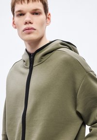 Mikina s kapucňou v olivovej zelenej farbe, s plným zipsom a čiernymi detailmi. Hladká textúra s pohodlným strihom.