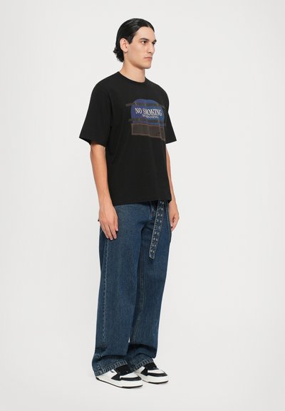 Camiseta de algodón negra con la impresión de "NO FUMAR" en azul y marrón en la parte delantera, combinada con pantalones de denim sueltos azules y zapatillas en blanco y negro.