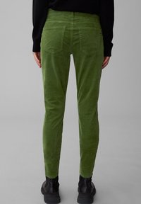 Persona con pantaloni di velluto a coste verde e stivali neri, in piedi con le mani rilassate ai lati su uno sfondo grigio uniforme.
