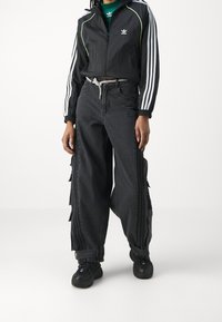 Svart atletisk jacka med vita ränder och gröna detaljer, i kombination med lösa svarta denimbyxor med sidodetaljer och dragsko. 