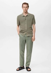 Hellolivgrünes, gestricktes Poloshirt mit kurzen Ärmeln, Knopfverschluss, strukturiertem Muster, kombiniert mit einer passenden, locker sitzenden Hose und schwarzen Sandalen.