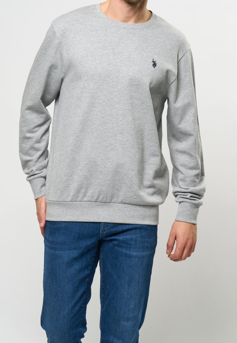 Gray cotton sweatshirt with a crew neck, long sleeves, and ribbed cuffs. Features a small navy logo on the chest. Paired with blue jeans.