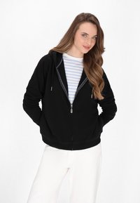 Schwarze Zip-up-Hoodie aus weichem Material, mit gefütterter Kapuze, Fronttaschen und silbernem Reißverschluss. Über einem hell gestreiften Shirt getragen.