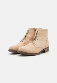 Bottes chevilles beiges en cuir texturé avec un bout arrondi, un laçage sur le devant et un petit talon en bois. Comprend une fermeture éclair latérale.