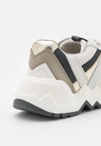 Talons de baskets épaisses en blanc, gris et or métallique avec semelle à motif zigzag et empiècements en mesh sur le côté.