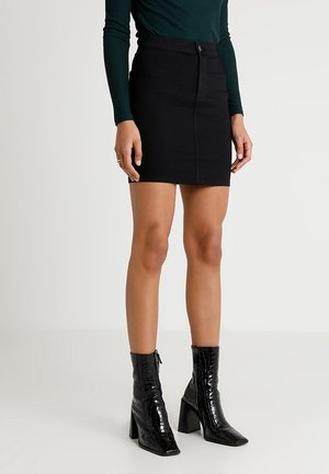 Mujer con una minifalda negra de talle alto, camiseta de manga larga verde oscuro y botines negros brillantes con tacón y textura de cocodrilo.