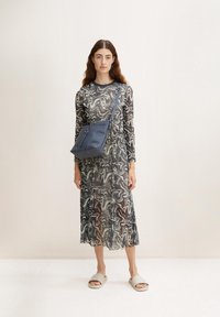 Abito lungo trasparente a motivi a spirale in bianco e nero, abbinato a una borsa a spalla blu e sandali leggeri grigio chiaro.