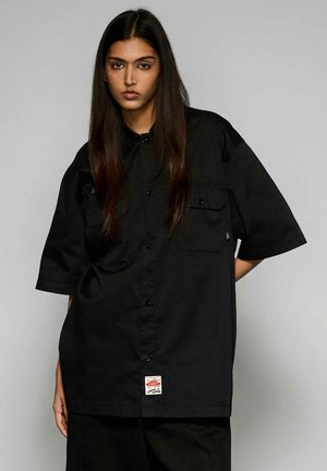 Jeune personne aux longs cheveux foncés portant une chemise noire boutonnée surdimensionnée avec des poches, sur un fond gris uni.