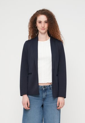 Junge Frau mit lockigem roten Haar trägt ein marineblaues Sakko, ein weißes, strukturiertes Croptop und blaue Jeans mit hoher Taille, steht vor einem schlichten Hintergrund.