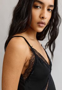 Fekete csipkével díszített felső, vékony pántokkal, V-kivágással és puha anyaggal, amelynek enyhe fényessége és finom bojt részletei vannak.