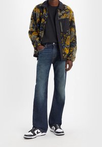 En man som bär en flerfärgad fleecejacka, svart skjorta, urblekta blå jeans och svart-vita sneakers, står med ena handen i fickan.