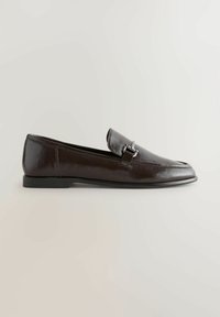 Mocassin en cuir marron foncé à enfiler avec un détail de boucle métallique argentée sur le dessus, semelle plate noire et bout rond lisse.
