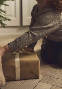 Una scatola regalo quadrata marrone avvolta in carta kraft, legata con un nastro crema, posata su un pavimento di legno. La mano di un bambino si allunga verso di essa.