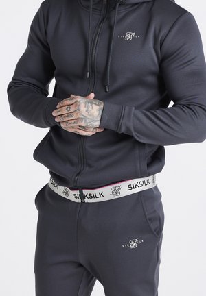 Mand iført sort SikSilk træningssæt med lynet hættetrøje og mærket taljebånd, hænder foldet og viser tatoveringer på fingre og knoer.