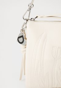 Borsa a tracolla in pelle bianca con design floreale in rilievo, hardware argentato e un charm a forma di cuore con una "D" nera. Chiusura con zip e tracolla.