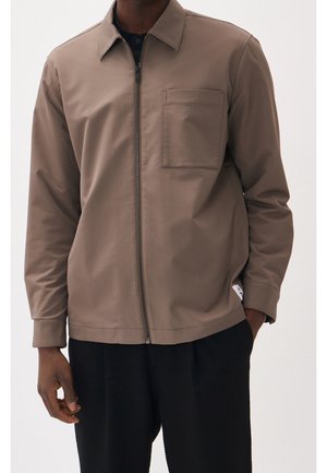 Hombre vestido con una chaqueta marrón con cremallera y bolsillo en el pecho delantero y pantalones negros, de pie con una mano en el bolsillo.