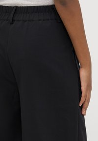 Black elastic-waist trousers with a loose fit, featuring smooth fabric and side pockets, partially visible with a hand resting on the side.