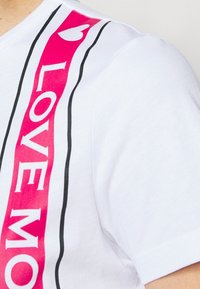Vit bomull t-shirt med en djärv rosa och svart "LOVE MORE" grafisk rand längs sidan och en liten hjärtaccent.