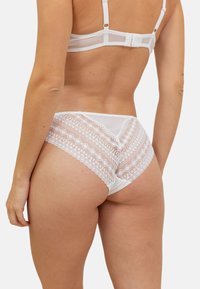 Culottes en dentelle blanche taille haute avec des motifs géométriques et des bords festonnés, présentant un corps en tissu lisse et un design arrière transparent.