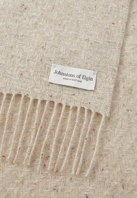 Kremasti šal od vune s uzorkom riblje kosti i resicama na rubovima. Na oznaci piše "Johnstons of Elgin, proizvedeno u Škotskoj." Meka tekstura, topao izgled.
