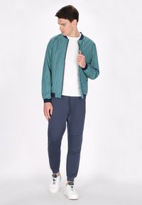 Teal bomberjack met geometrisch patroon, marineblauwe afwerking; witte t-shirt, marineblauwe joggers en witte sneakers. Glad stof met gestructureerd ontwerp.