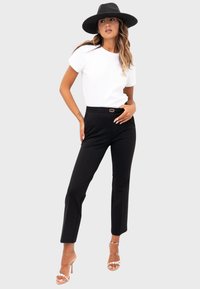 Zwarte tapered broeken met een gouden accent op de tailleband, gecombineerd met een aansluitend wit t-shirt en transparante hakken, gestyled met een hoed met brede rand.