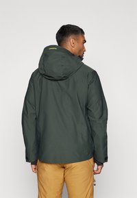 Chaqueta impermeable verde con capucha, que presenta una textura suave, puños elásticos y un detalle de logo amarillo en contraste en la parte posterior.