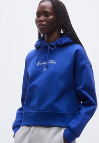 Modrá bavlnená mikina s kapucňou naťahovacou šnúrkou a vyšívaným logom "Calvin Klein". Mäkká textúra a voľný strih, so žebrovanými manžetami a pásom.