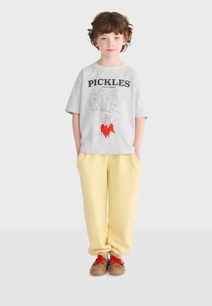 Enfant aux cheveux bruns bouclés, debout face à l'avant, portant un t-shirt gris avec le texte "PICKLES", un pantalon jaune et des chaussures marron avec des lacets rouges.