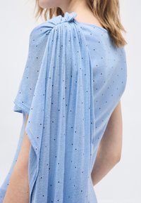 Γυναίκα που φοράει τοπ από ανοιχτό γαλάζιο ύφασμα με μικρές μαύρες κουκκίδες και λεπτομέρεια με πτυχώσεις και ραφή στα μανίκια στην περιοχή του ώμου.