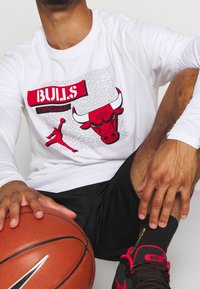 Camisa blanca de manga larga con el texto "BULLS" en rojo y un gráfico de un toro en negro y rojo, combinada con pantalones cortos negros y calzado negro de Nike. Incluye un balón de baloncesto.