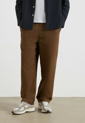 Braune, locker sitzende Hosen aus weichem Stoff, kombiniert mit grauen und weißen Turnschuhen. Das Modell trägt ein schwarzes Hemd über einem weißen T-Shirt.