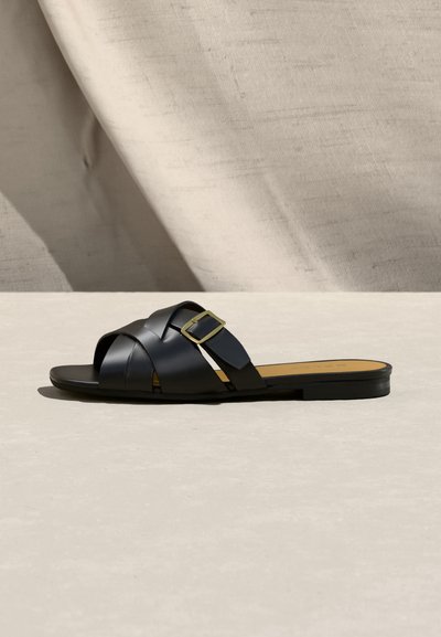 Sandale plate en cuir noir avec brides croisées et boucle dorée sur une surface beige avec un fond en tissu neutre.