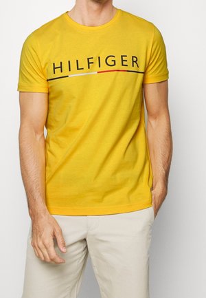 Mann trägt ein gelbes kurzärmeliges T-Shirt mit dem Schriftzug "HILFIGER" und Farbblock-Linie, kombiniert mit hellbeigen Hosen.