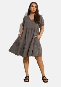 Vestido gris de capas con mangas cortas, escote en V, bolsillos laterales, hecho de una tela ligera, combinado con sandalias negras con puntos.