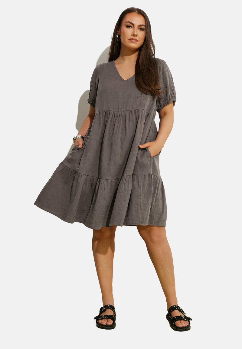 Vestido gris de capas con mangas cortas, escote en V, bolsillos laterales, hecho de una tela ligera, combinado con sandalias negras con puntos.