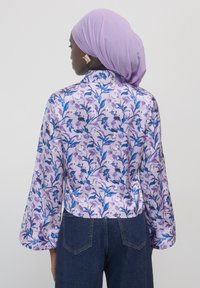 Langärmlige Bluse aus leicht lila Satin mit floralem Muster in Blau und Lila. Kombiniert mit dunklen Jeans, zeigt die Rückansicht.