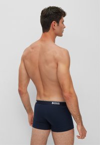 Marineblaue Boxershorts aus einem glatten, dehnbaren Stoff mit einem kontrastierenden weißen "BOSS"-Logo am Bund. Modell von hinten gezeigt.