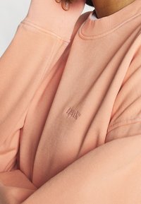 Sudadera de algodón rosa con cuello redondo y puños acanalados, que presenta un sutil logo tonal bordado en el pecho. Textura suave.
