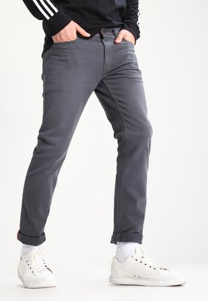 Man met opgerolde grijze broek, witte sokken, witte sneakers en een zwart shirt met lange mouwen met witte strepen, staand met handen in de zakken.