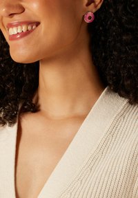 Ronde oorbellen met roze emaille en gouden accenten, met een donutontwerp, gedragen door een persoon met krullend haar en een neutrale trui.