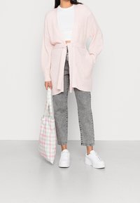 Svetle růžový kardigan s otevřeným úpletem a zavazováním v pase, nošený přes bílý crop top, doplněný šedými džíny s vysokým pasem a bílými teniskami.