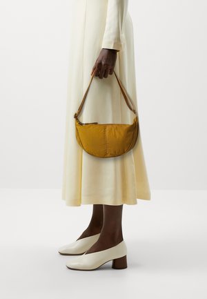 Personne en robe crème tenant un sac à main jaune moutarde à une seule bandoulière, portant des chaussures blanches à talons blocs sur un fond uni.