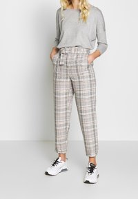 Szara bluzka z długim rękawem w połączeniu z spodniami w kratę o wysokim stanie w kolorze beżowym, czarnym i pomarańczowym. Spodnie mają kieszenie boczne i przewiązany pasek. Białe sneakersy.