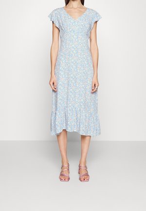 Femme portant une robe midi fleurie bleu clair avec des manches papillotes et des sandales roses à talons blocs avec brides, debout devant un fond uni.