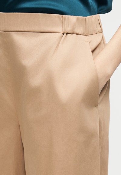 Άτομο με μπεζ παντελόνι, το χέρι στην δεξιά τσέπη, και μπλε-πράσινο πουκάμισο μαζεμένο στη μέση.